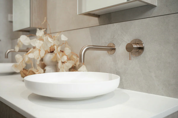 Modern brushed brass basin mixer mounted above a white round countertop basin in a contemporary bathroom